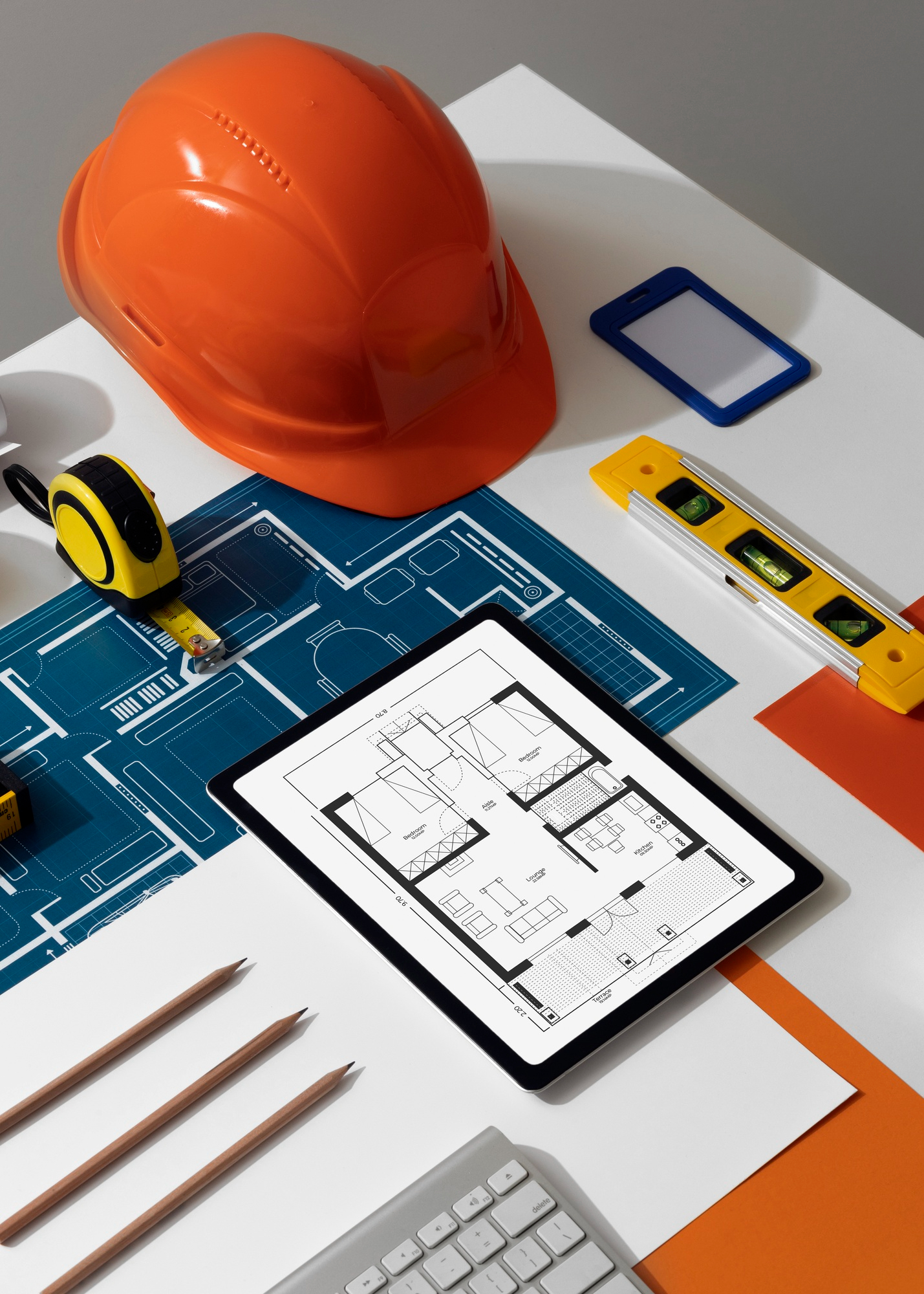Ujęcie z góry na biurko budowlane ze sprzętem i planami. Centralnym punktem jest tablet wyświetlający czarno-biały rzut architektoniczny mieszkania. Wokół leży pomarańczowy kask ochronny, niebieska kalka techniczna z planami, żółta poziomica, miarka budowlana i trzy ołówki. Częściowo widoczna jest klawiatura komputerowa.