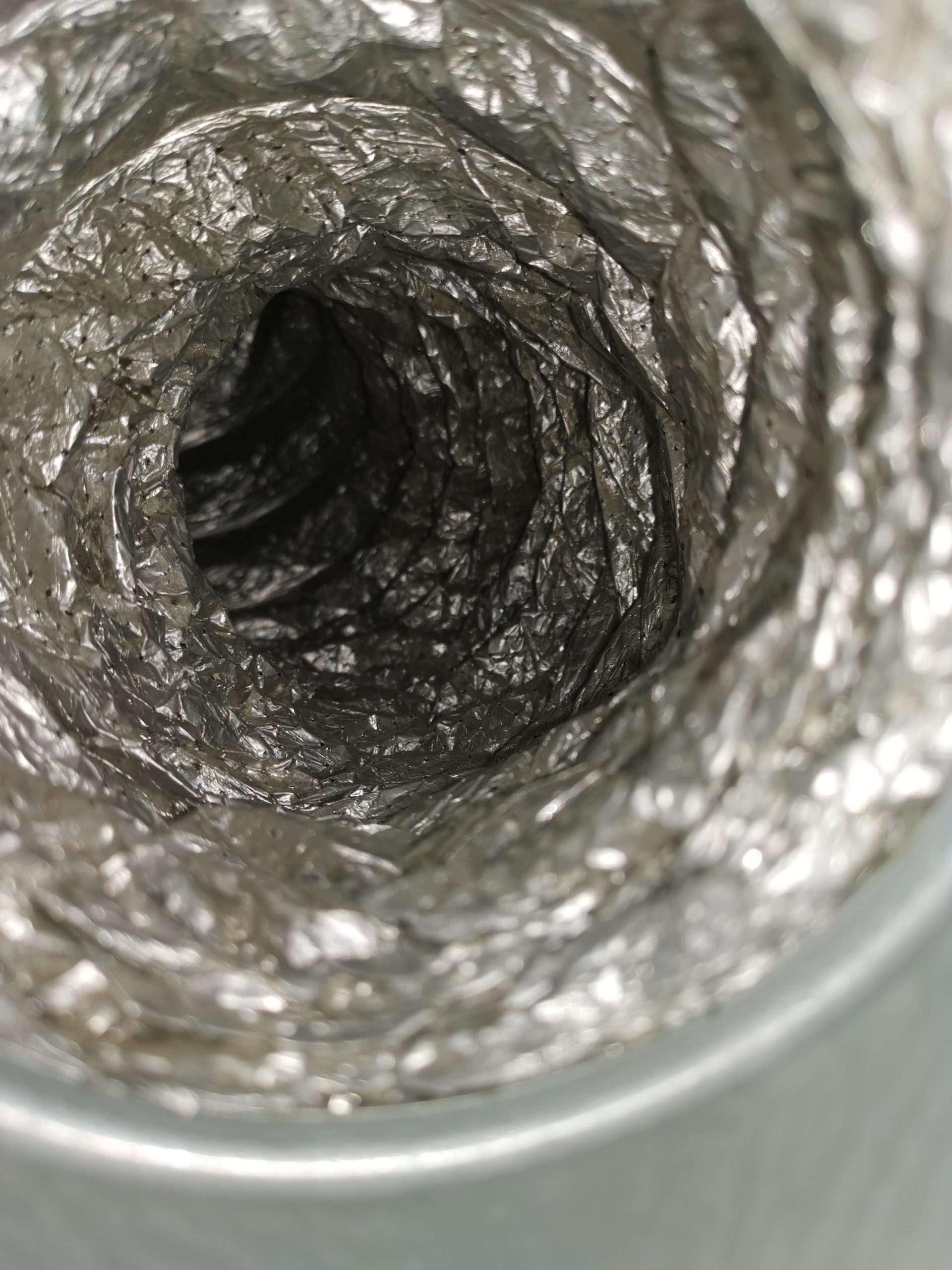 Zbliżenie na wnętrze karbowanego, elastycznego przewodu wentylacyjnego (kanału spiro), wykonanego z folii aluminiowej. Widoczna jest błyszcząca, pognieciona tekstura ścianek przewodu, tworząca głęboki, ciemny tunel.